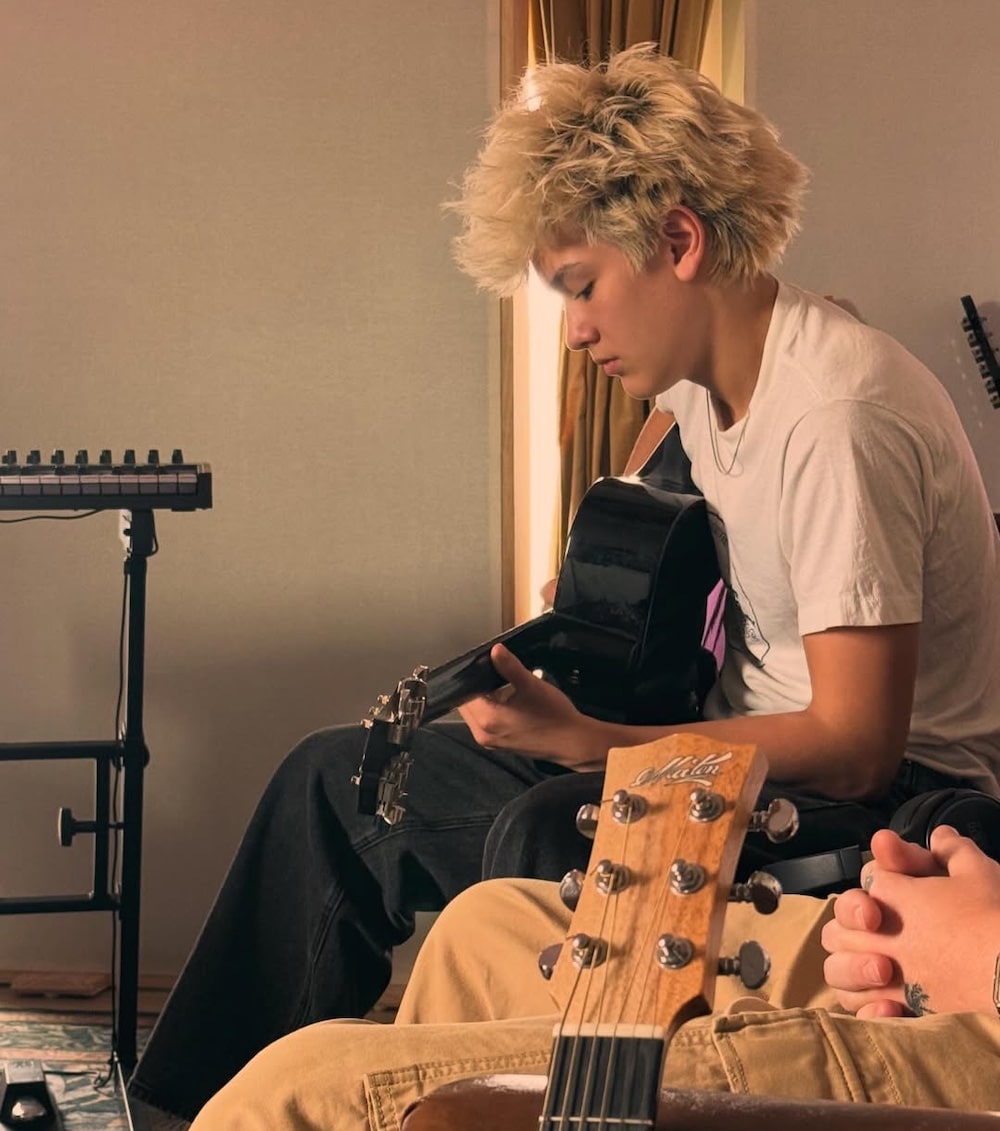 A person with blond, tousled hair sits on a couch playing a black acoustic guitar. Another guitar and a keyboard are visible nearby, creating a relaxed vibe reminiscent of a Girls' Life June/July issue music session.