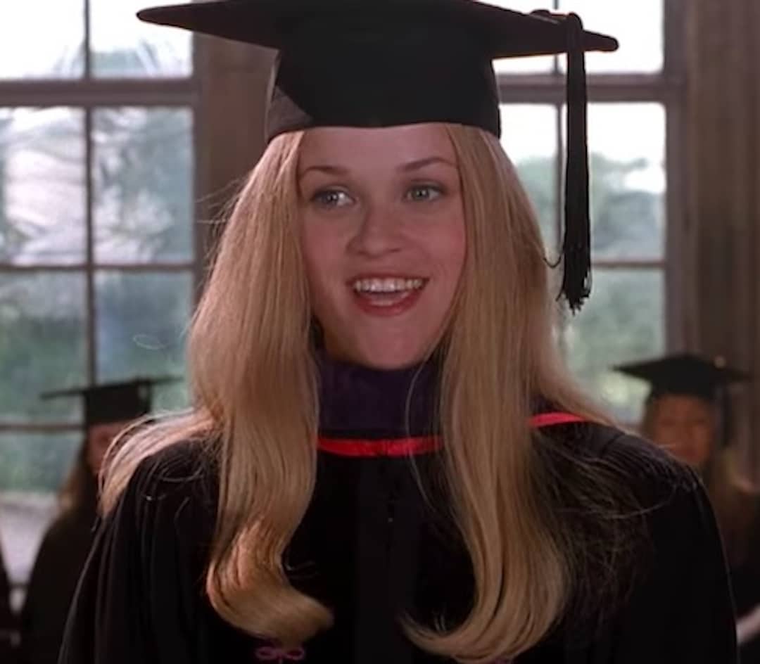 A smiling woman with long blonde hair, wearing a black graduation cap and gown with a purple and red trim, stands indoors in front of large windows, celebrating her graduation goal as other graduates are visible in the blurred background.