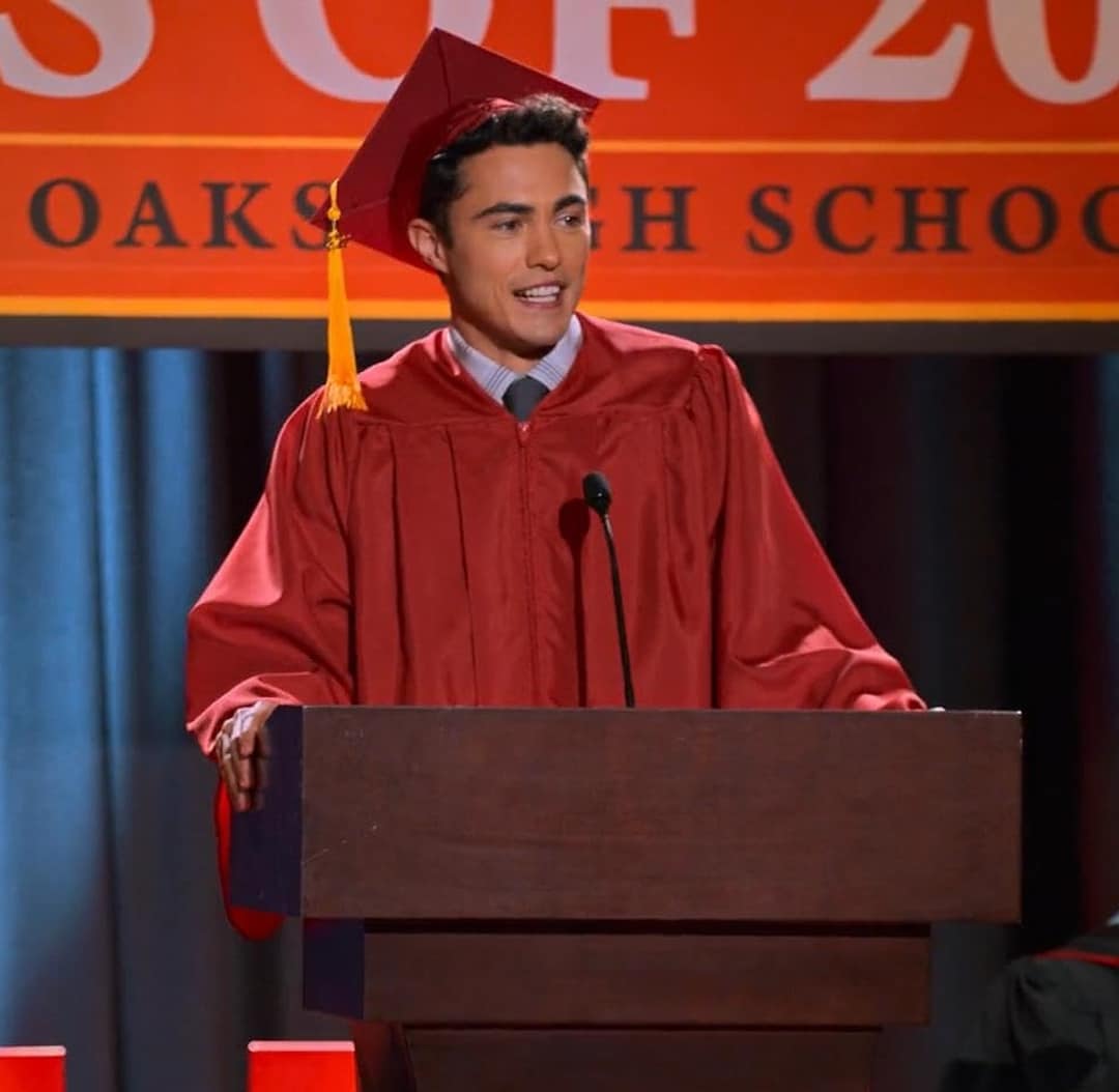 A young man in a red graduation cap and gown stands at a podium, speaking about his graduation goal. Behind him is a sign that partially reads OAKS HIGH SCHOOL and CLASS OF. He appears to be delivering an inspiring graduation speech.