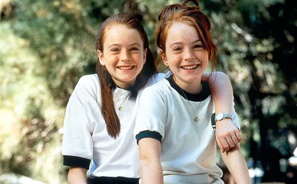 Two young girls with long red hair, wearing casual shirts, sit closely together outdoors, smiling at the camera. One girl has her arm around the other's shoulders. Green trees are blurred in the background for a June/July issue of Girls' Life.