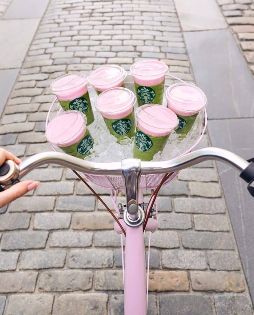 A person rides a pink bicycle with a basket filled with ice and seven Starbucks cups with pink lids, delivering the perfect summer order on a cobblestone street.