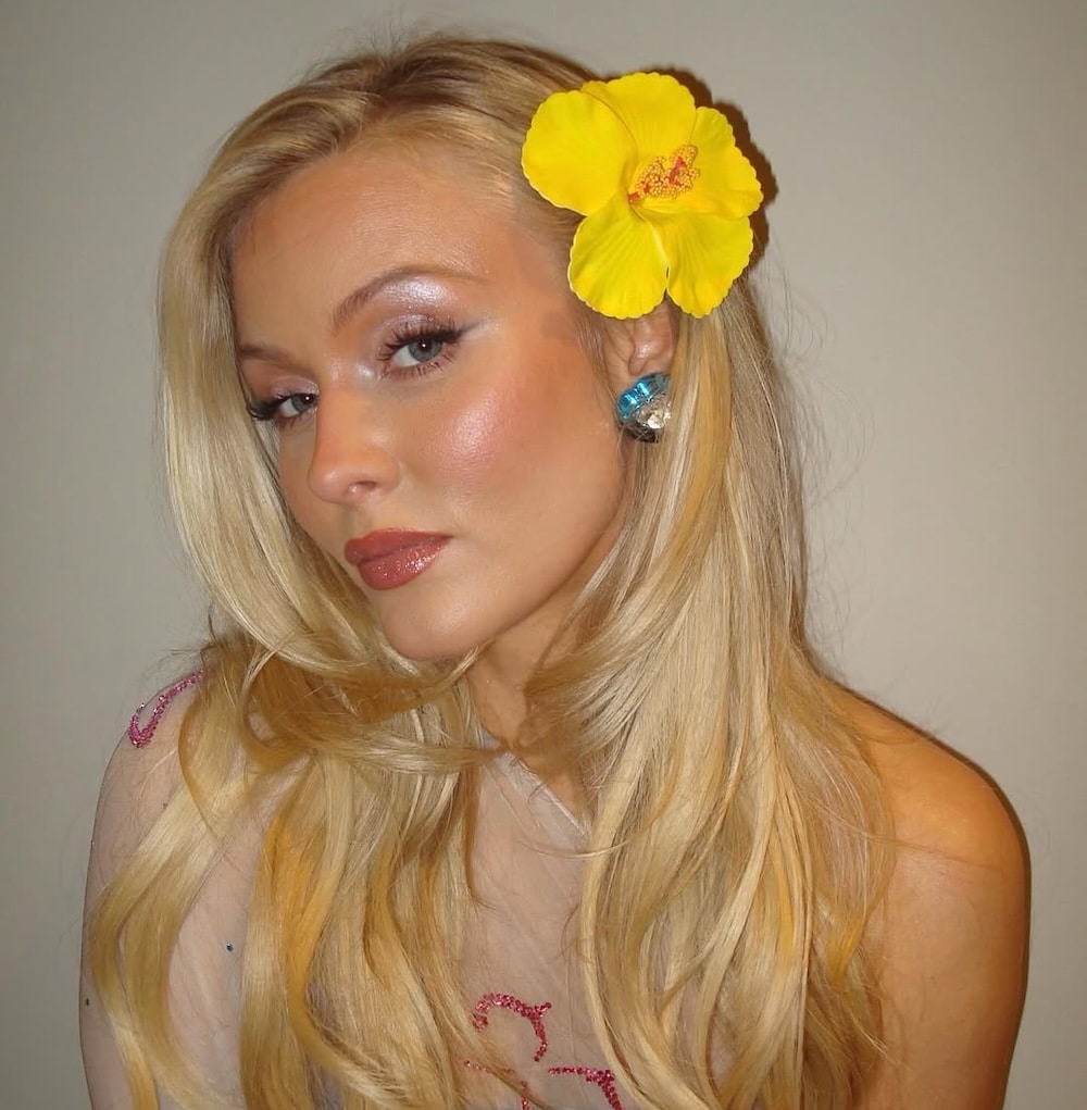 A woman with long blonde hair adorned with a large yellow flower above her ear poses against a plain background, channeling Girls' Life June/July issue vibes. She sports shimmery makeup, silver and turquoise earrings, and a sheer top with pink details.