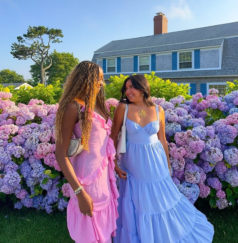 Two women in colorful, flowing dresses stand smiling in front of blooming hydrangeas, capturing a scene reminiscent of a Girls' Life June/July issue. One woman wears pink, the other light blue, with a house and blue sky behind them.