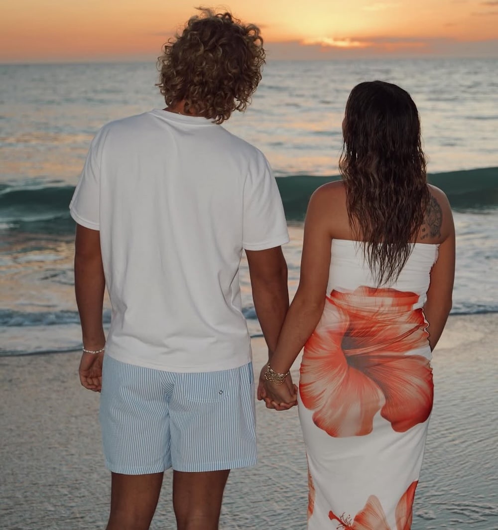 A couple stands hand in hand on the beach, facing the ocean at sunset, channeling breezy June/July issue vibes. The man wears a white T-shirt and striped shorts, while the woman’s white dress features a bold red flower print.