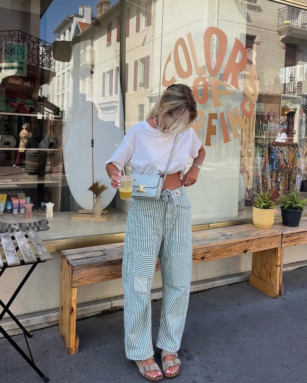 A woman stands on a sidewalk in front of a shop window, wearing a white t-shirt, striped wide-leg pants, sandals, and a light blue crossbody bag—channeling Malia Baker’s chic vibe from the June/July issue of Girls' Life—as she sips her drink and looks down.