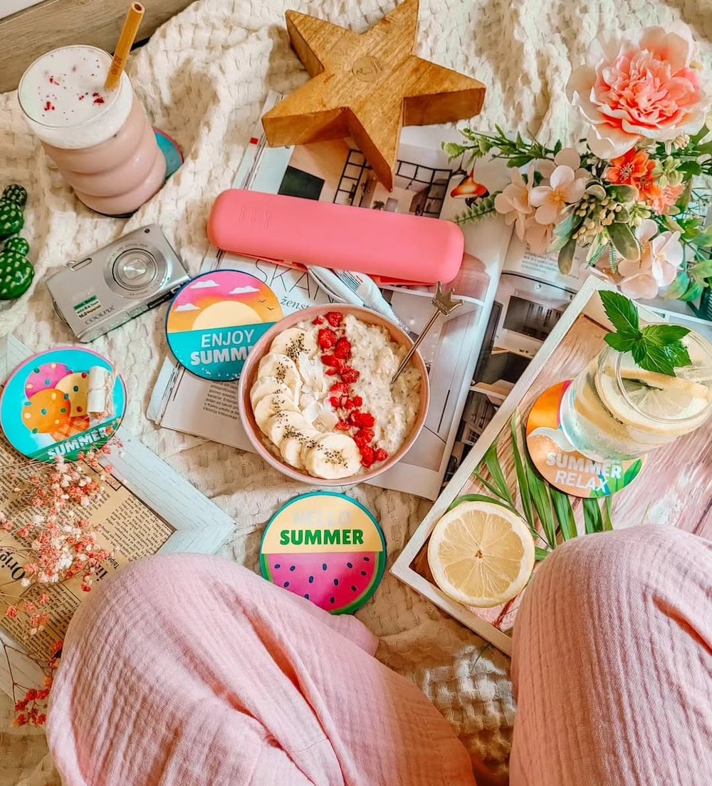 A cozy summer picnic setup on a blanket features a smoothie, oatmeal with banana and berries, lemon water, flowers, magazines including the Girls' Life June/July issue starring Malia Baker, colorful coasters, a camera, and someone in pink pants.
