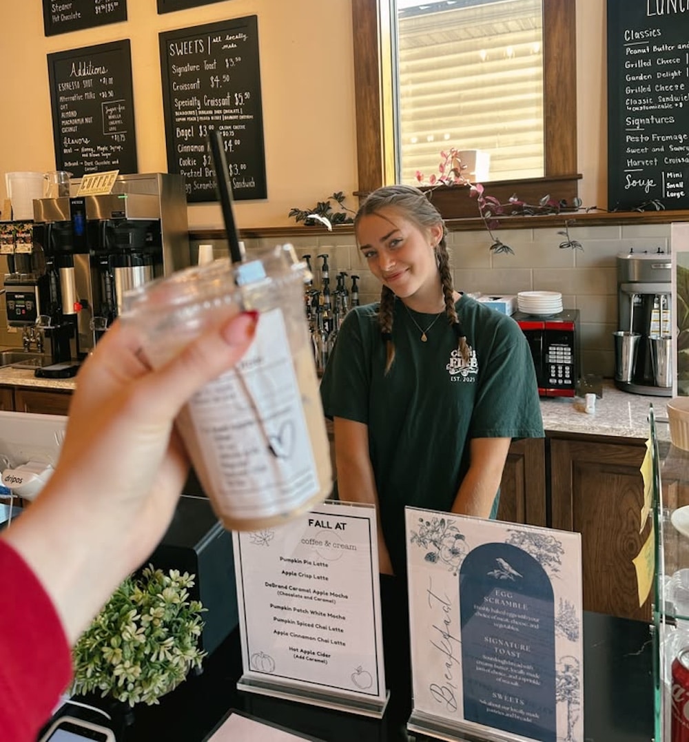 A person holds up an iced coffee in the foreground, while Malia Baker, featured in the June/July issue of Girls' Life, smiles behind the counter of a cozy café with menu boards on the wall.