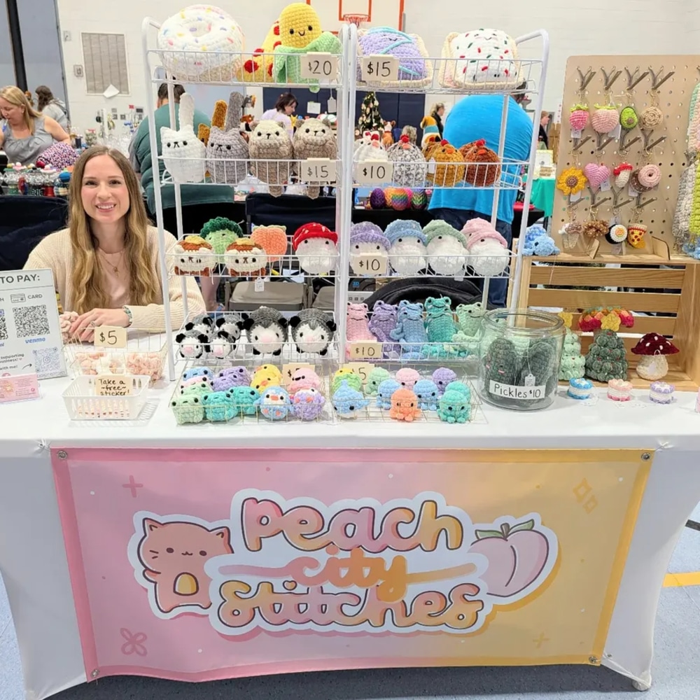 A smiling woman sits behind a booth displaying colorful crocheted plushies shaped like animals and food. A sign reads Peach City Stitches—a cheerful money-making summer setup with prices and payment info sheet on the table.