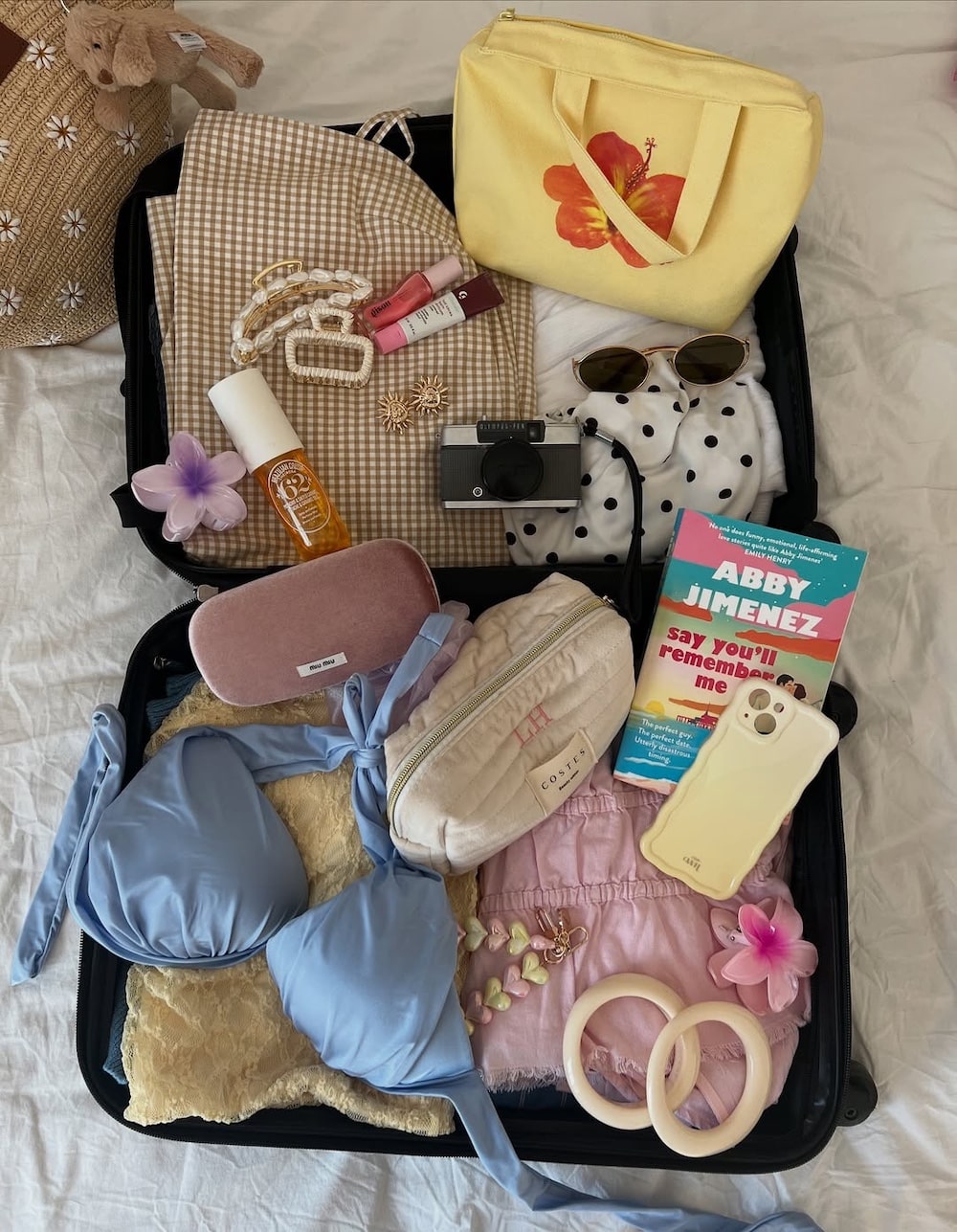 Open suitcase with summer items—blue bikini, pink shorts, polka dot shirt, camera, book, sunglasses, yellow bag and more—arranged neatly on a white bed with pink flower decorations for a Girls' Life June/July issue featuring Malia Baker.