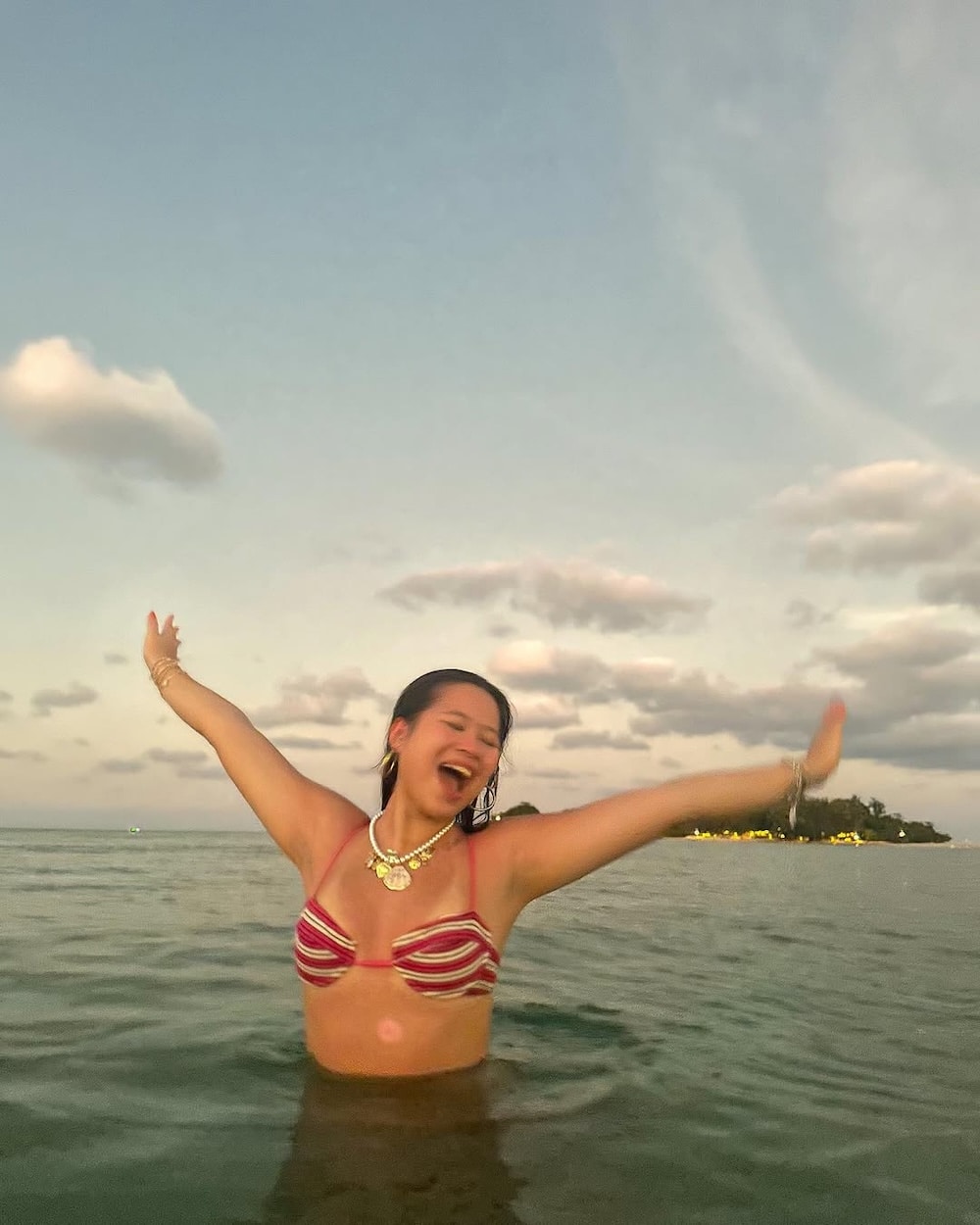 A joyful woman, reminiscent of Girls' Life's June/July issue, stands in the ocean with arms wide open, smiling as the sun sets. The sky is partly cloudy, and a distant shoreline with trees is visible in the background.