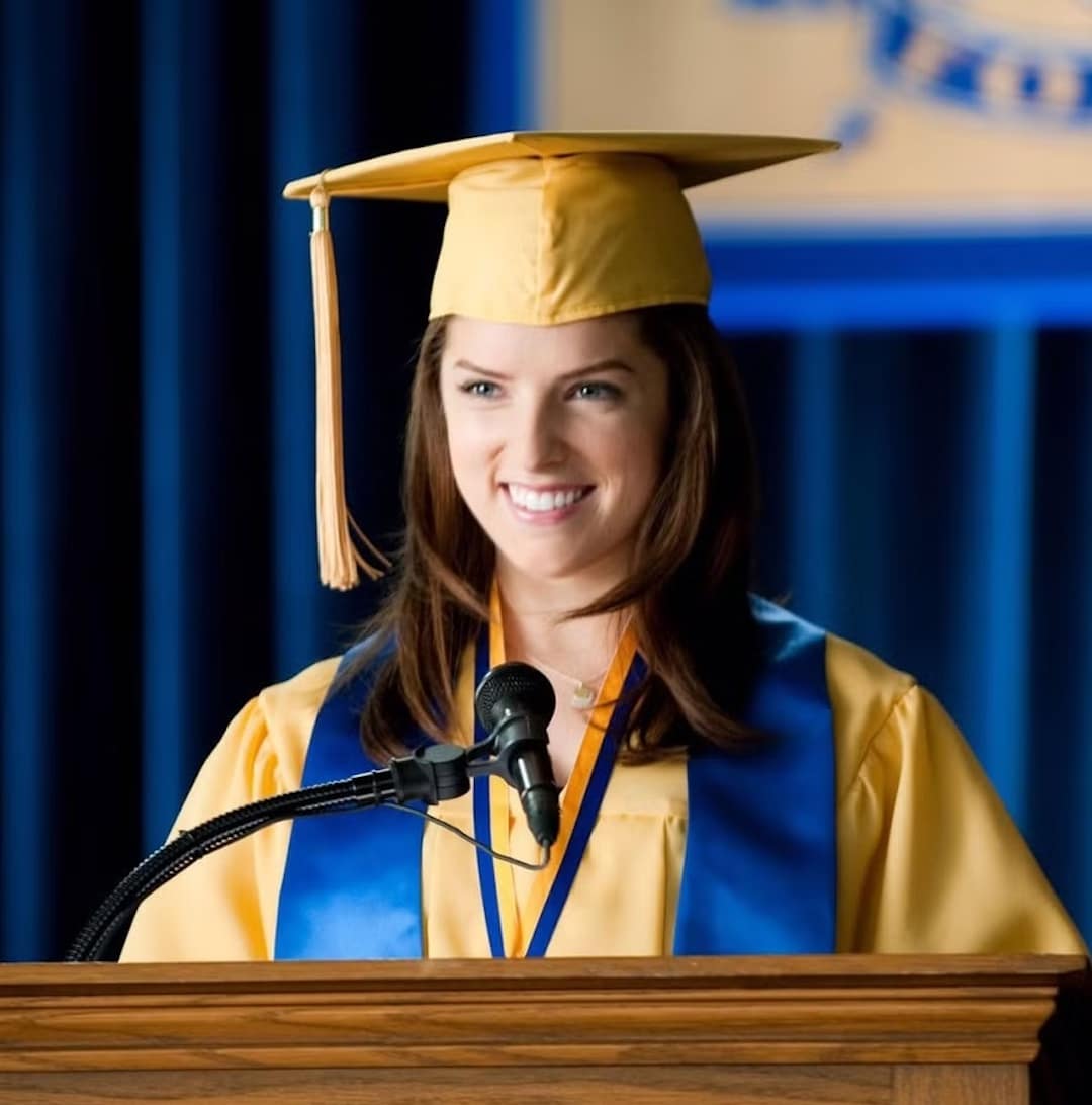 A young woman in a yellow graduation cap and gown with a blue stole smiles while standing at a podium, sharing her graduation goal into the microphone at a graduation ceremony.