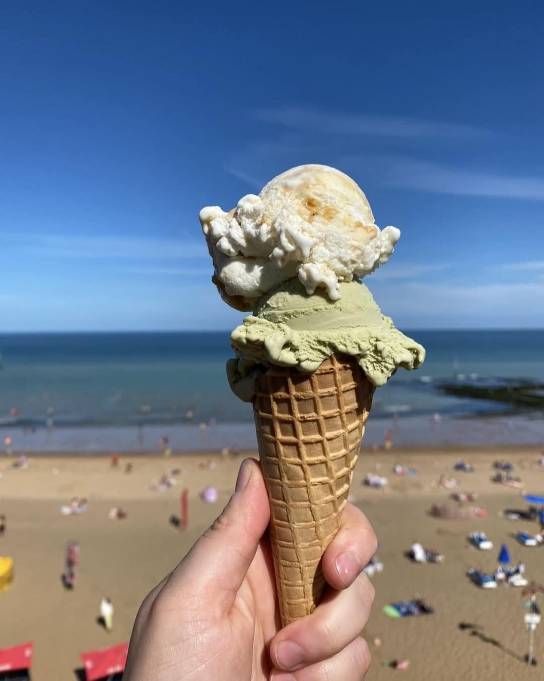 A hand holds an ice cream cone with two scoops—one green and one white—against the backdrop of a sunny beach with people lounging on the sand and the sea in the distance.