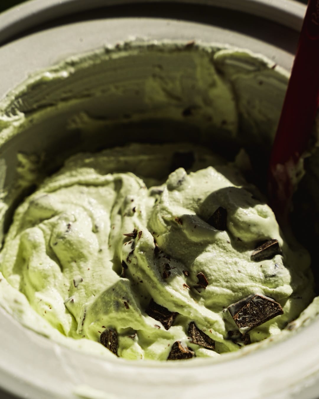 A close-up of mint chocolate chip ice cream being mixed in an ice cream maker, showing creamy pale green ice cream with chunks of chocolate throughout.