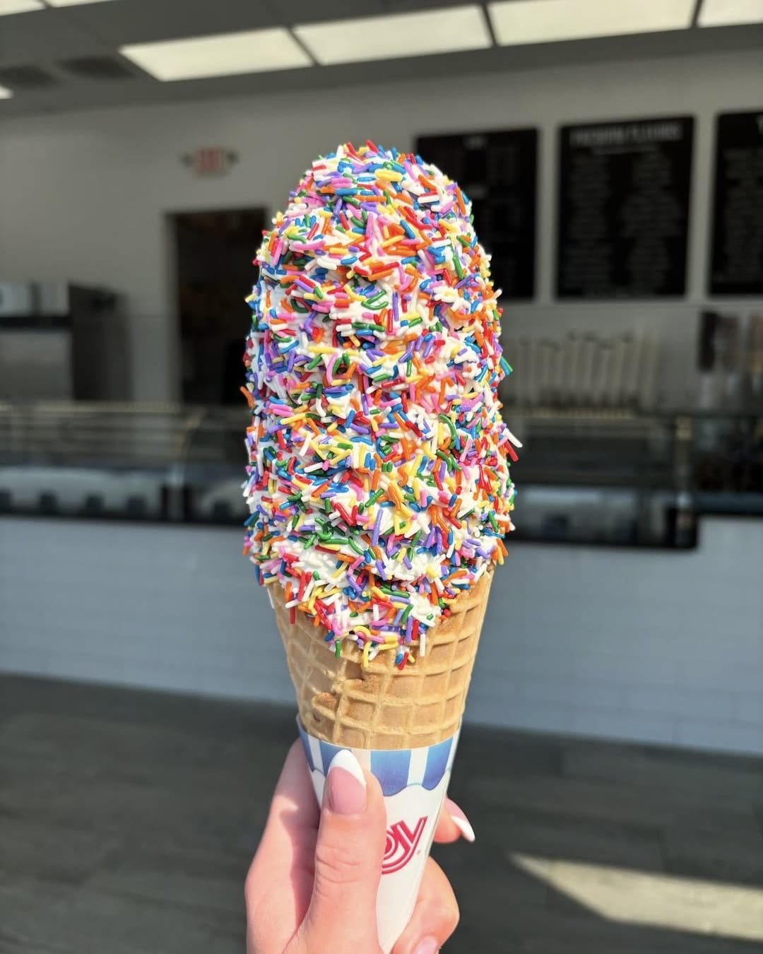 A hand holds a large ice cream cone covered in colorful rainbow sprinkles, set against the background of a bright ice cream shop with white tiles and menu boards.