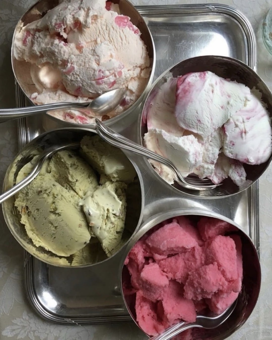 Four metal bowls filled with different flavors of ice cream or sorbet—pink, green, white with pink swirls, and white with red bits—sit on a silver tray. Each bowl has a metal spoon inside.