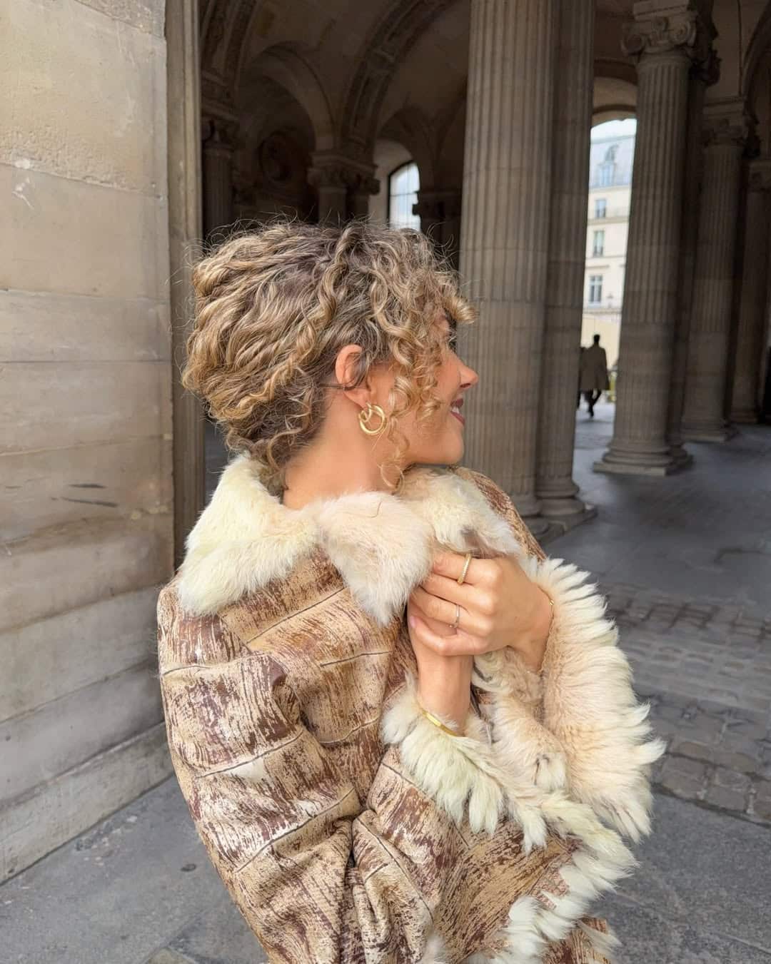 A woman with curly hair stands in a stone archway, wearing a fur-trimmed coat and gold hoop earrings. She is smiling and looking to the side, holding the coat close around her. Tall columns and soft light complete the elegant scene.
