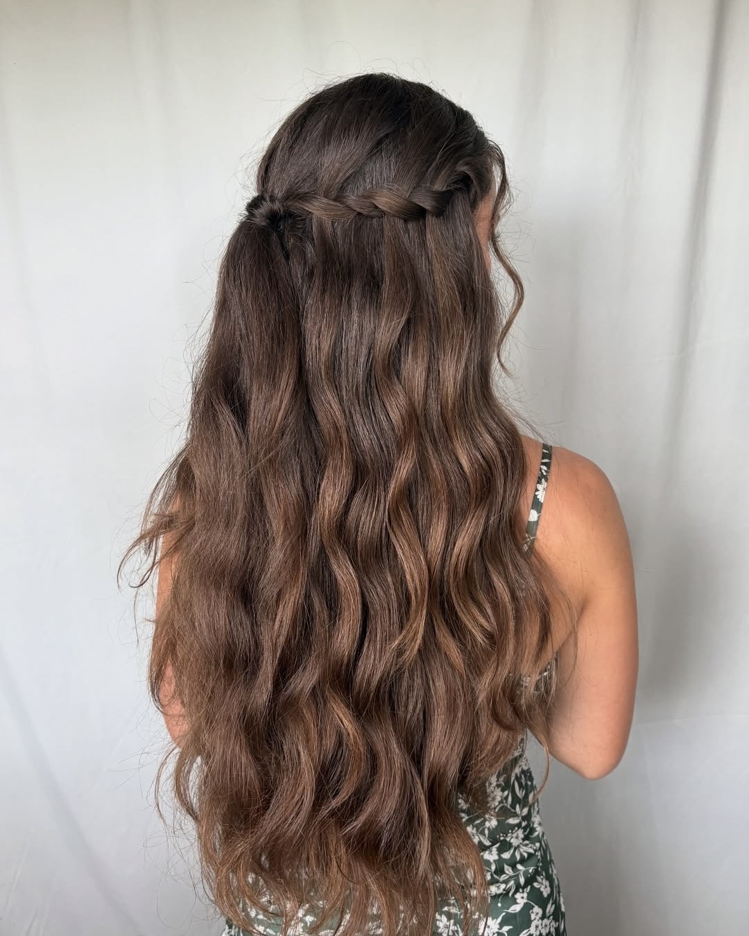 A woman with long, wavy brown hair styled in a half-up braid stands facing away, wearing a green, floral-patterned dress against a plain light backdrop.