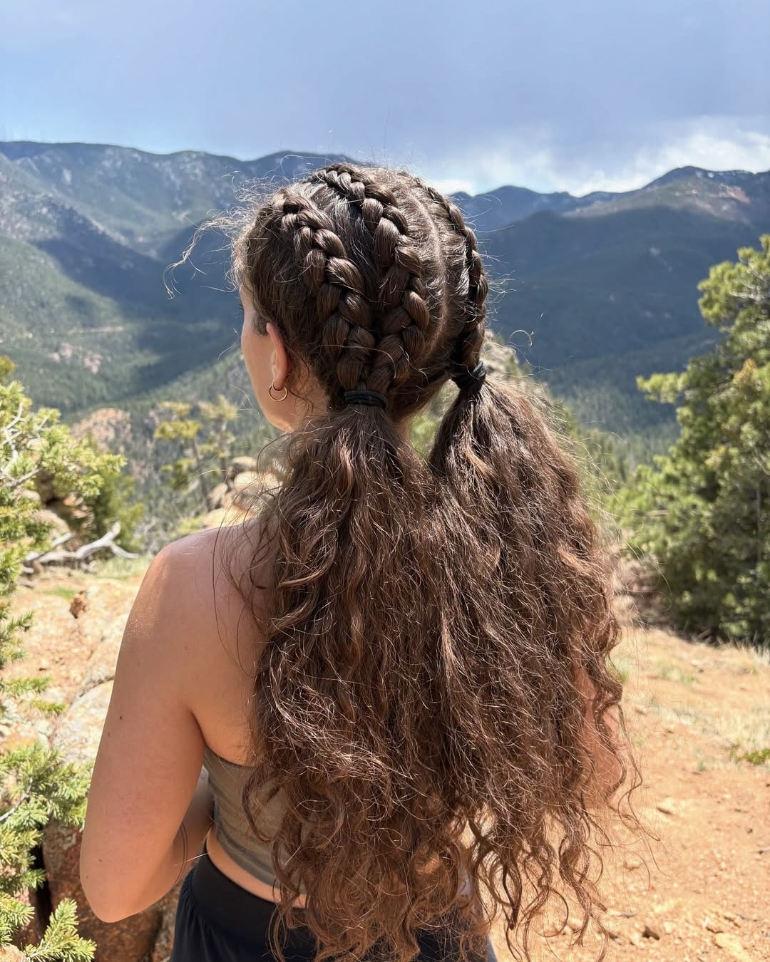 A woman with long, curly hair styled in braids stands outdoors, facing a scenic mountain landscape under a partly cloudy sky. She wears a tank top and is surrounded by greenery and rocky terrain.