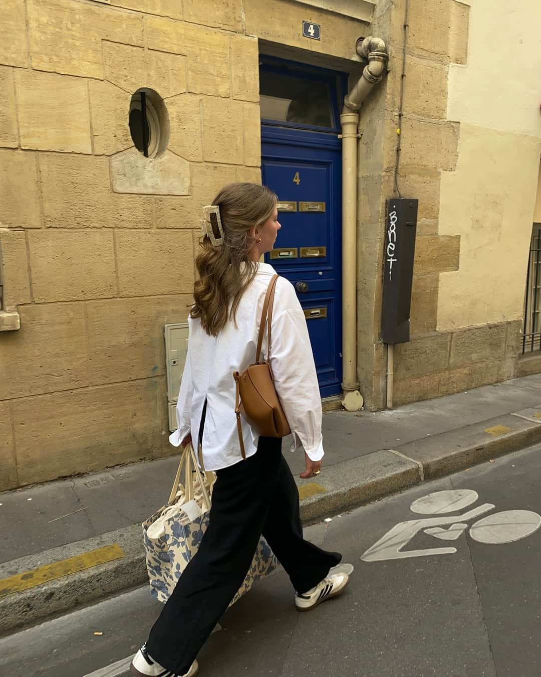 A woman with wavy hair, wearing a white shirt, black pants, and white sneakers, walks on a Paris street carrying a floral tote bag—ready to level up her style as if updating her resume for senior year. She passes a blue door and beige stone wall.