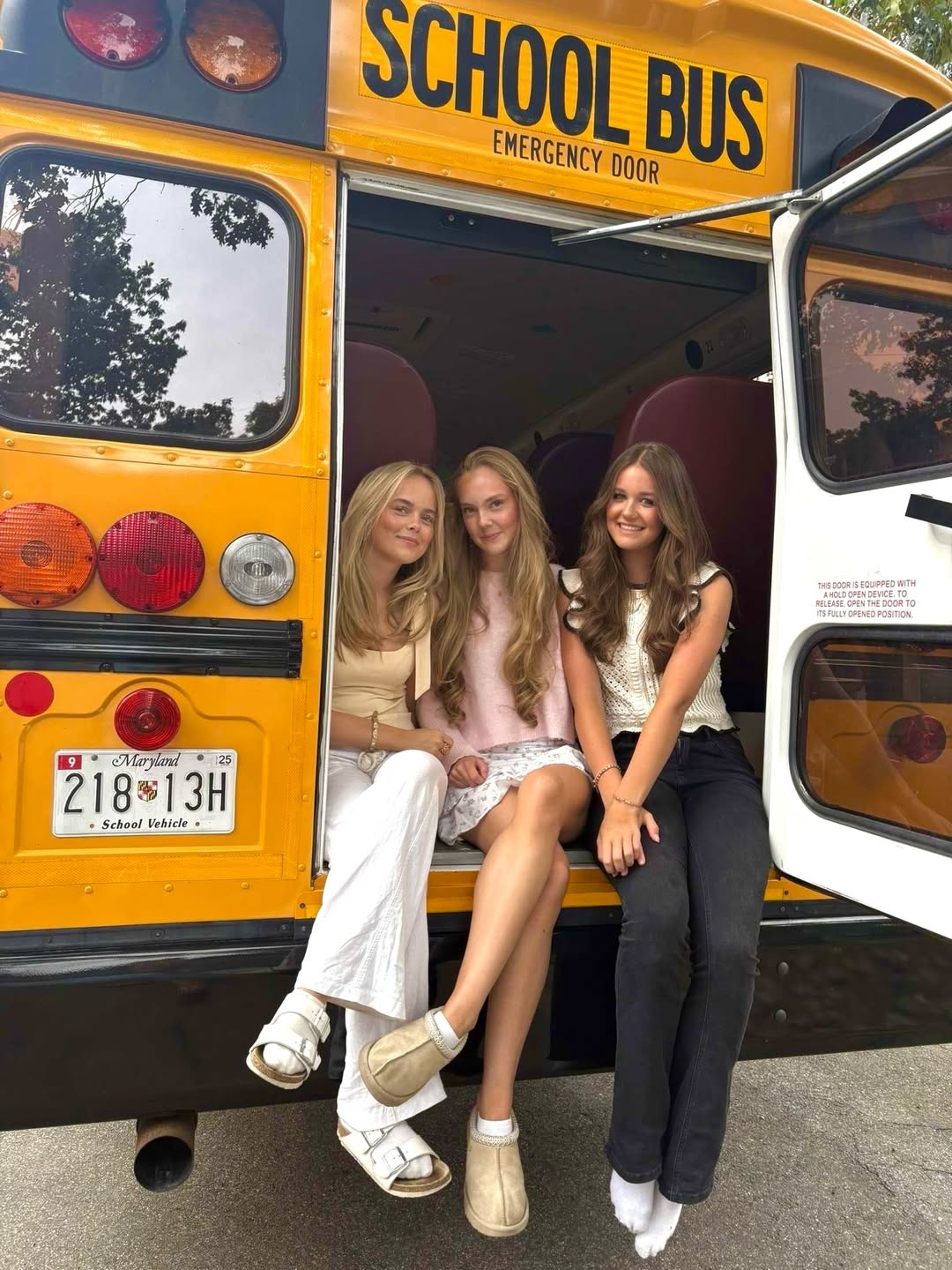 Three young women sit on the open back of a yellow school bus, smiling at the camera as they level up for senior year. The Maryland license plate and open emergency door are visible, with trees reflected in the bus windows.