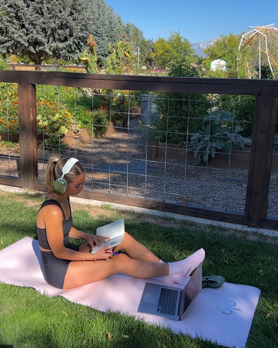 A woman sits on a yoga mat outdoors, wearing headphones and athletic wear, reading a book with "Level Up Your Resume" tips on her laptop. She enjoys the grassy area near a garden and wooden fence under clear blue skies.