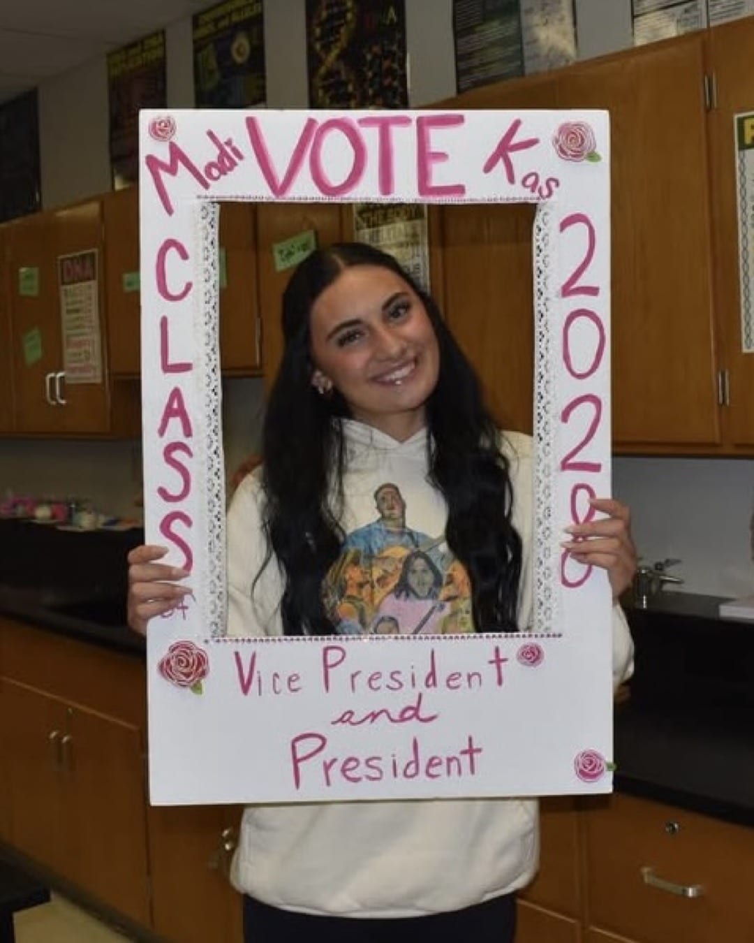 A smiling student holds a decorated frame reading Madi VOTE Kas, Class 2026, Vice President and President—showing how to level up your resume with leadership roles—in a classroom with posters and wooden cabinets in the background.