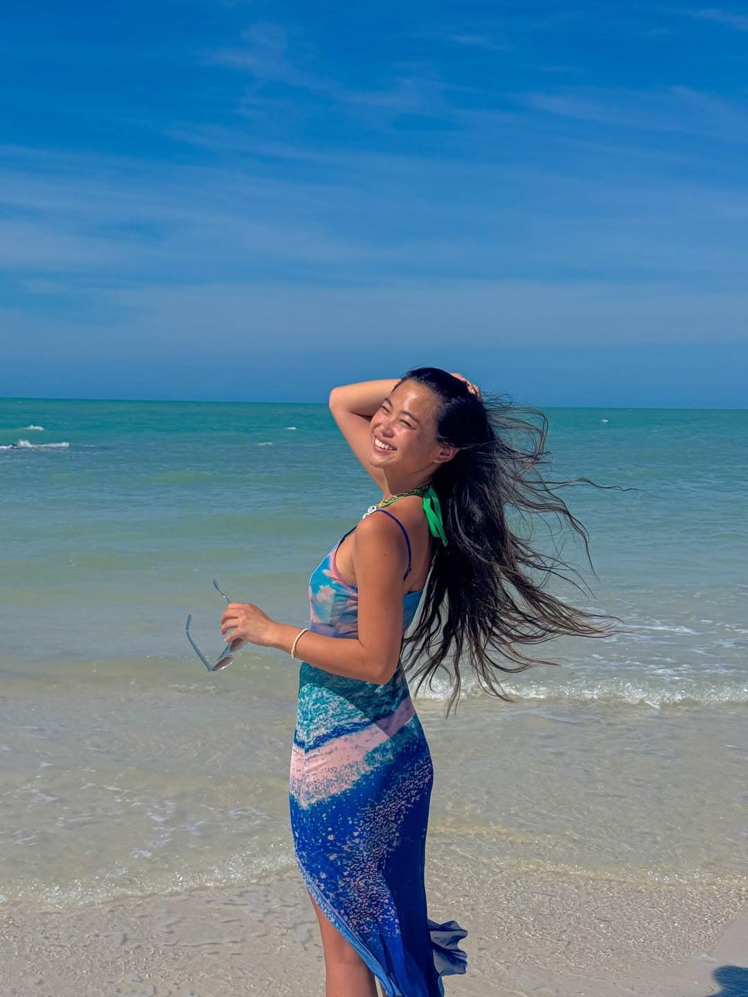 A woman in a blue dress stands on a sandy beach with the ocean behind her, smiling and holding sunglasses as her long hair blows in the wind under a clear blue sky.