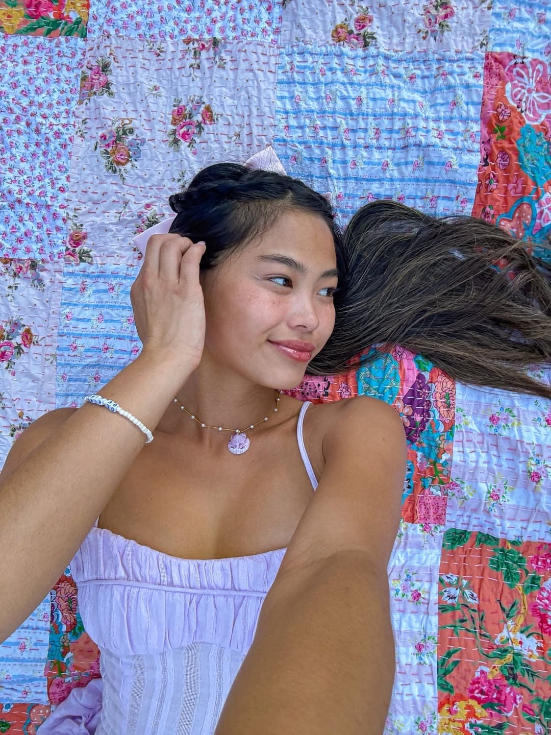 A woman in a light purple dress lies on a colorful patchwork quilt, smiling slightly as she touches her hair. Her long brown hair is spread out, and she wears a necklace and bracelet.