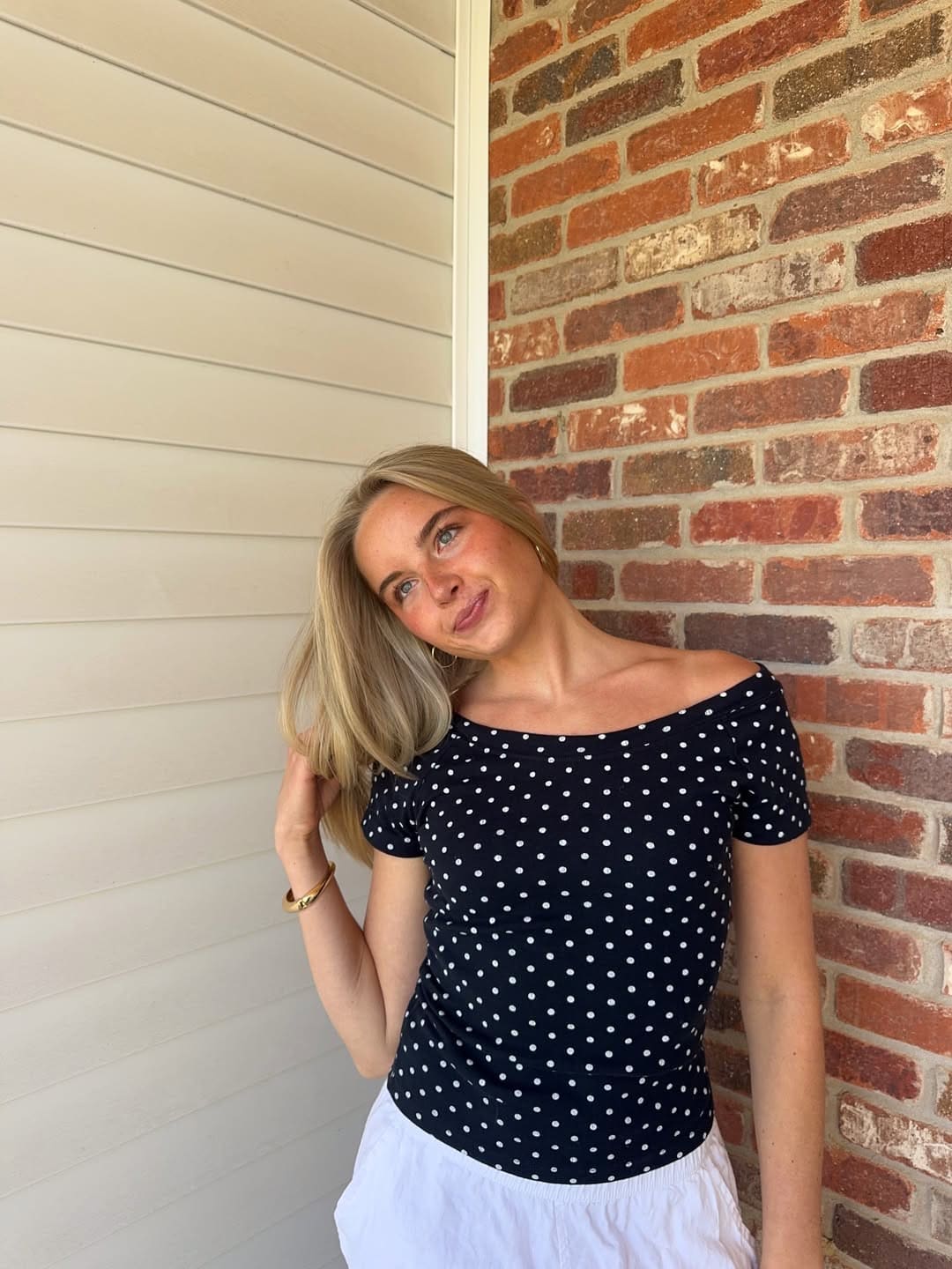A woman with long blonde hair, wearing a black and white polka dot top and white skirt, stands in front of a wall with vertical white paneling and red brick, smiling and touching her hair.