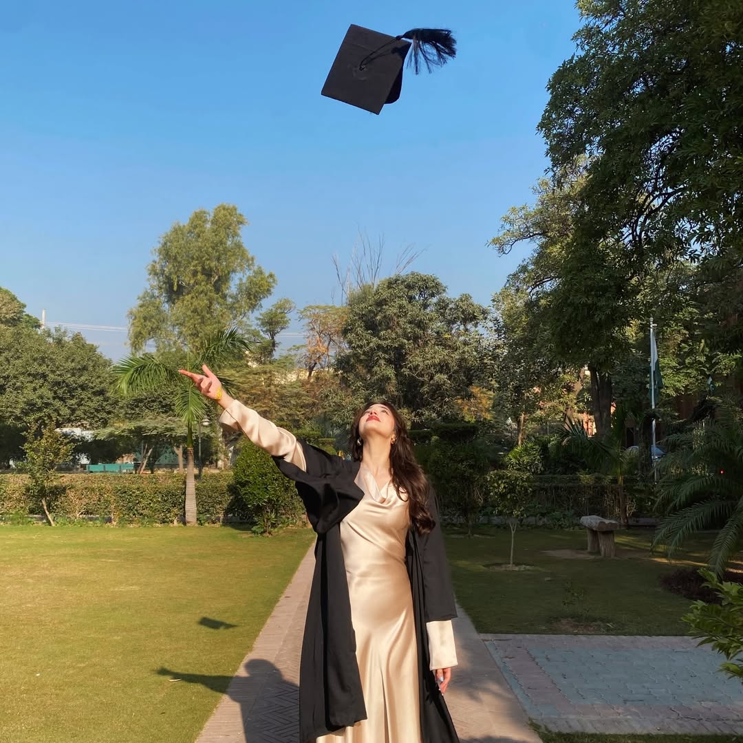 A woman in a cream dress and black graduation gown stands outside on a sunny day, smiling and tossing her cap into the air to celebrate the end of the school year, surrounded by greenery and trees.