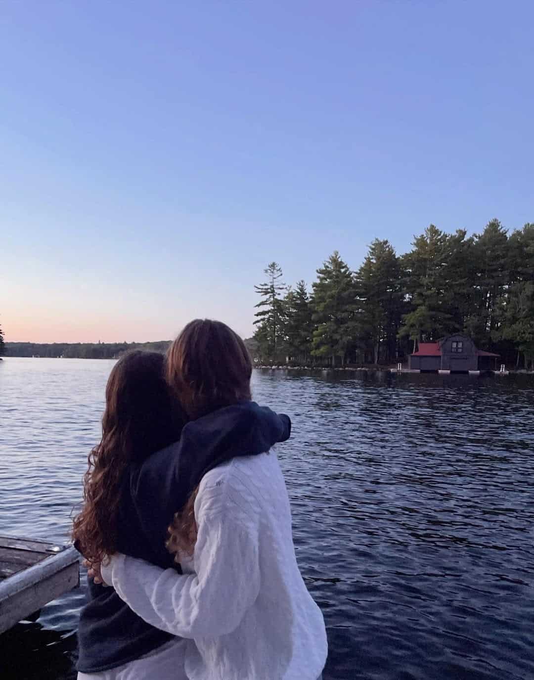 Two people with long hair stand on a dock by a lake, sharing a cute friend gesture as they hug and gaze at the sunset over the water and trees, creating a peaceful and tranquil scene.