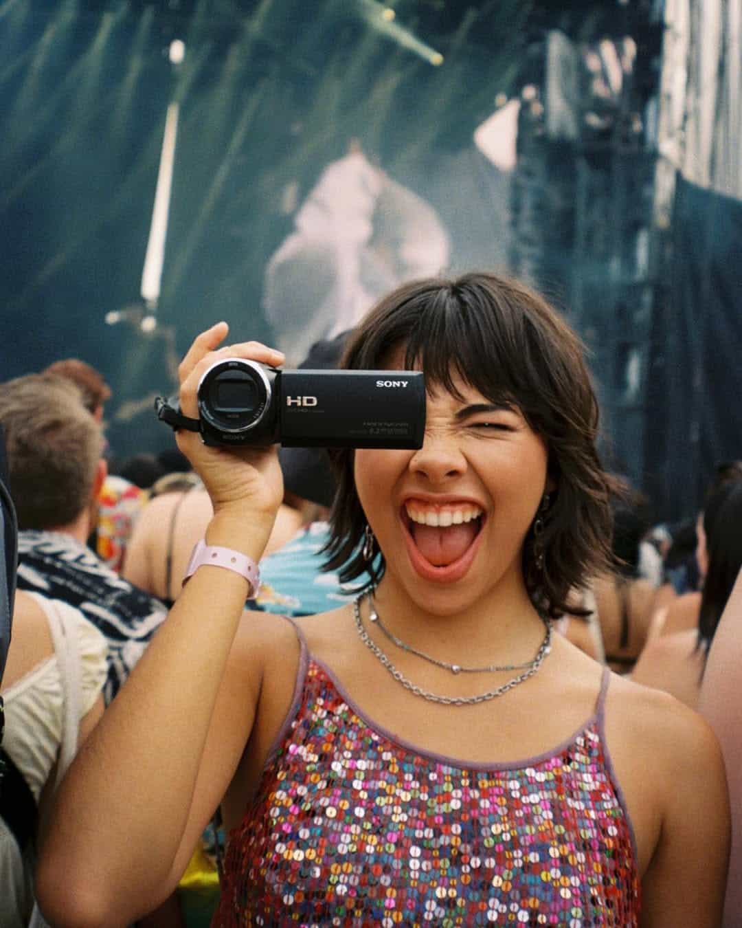A young woman in a sequined tank top smiles widely and winks while holding an HD Sony camcorder, recording at an outdoor concert or festival, surrounded by a crowd.