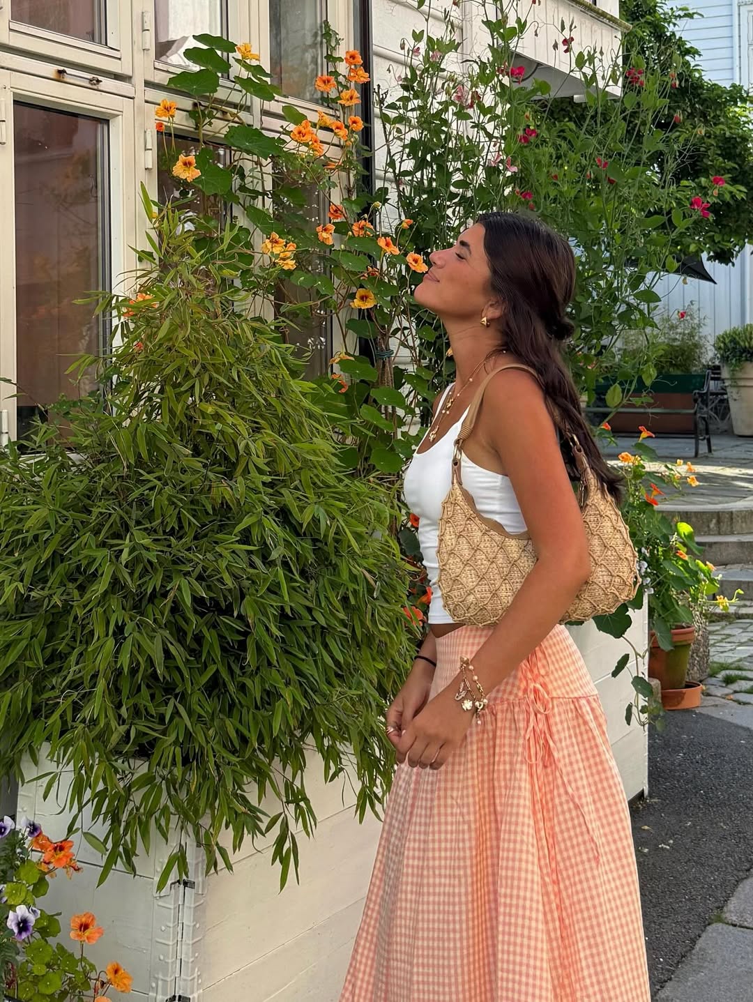 A woman in a white top and pink checkered skirt stands by a window, surrounded by green plants and orange flowers. With her woven bag and effortless style, she captures that cool girl summer vibe while enjoying a beautiful garden on a budget.