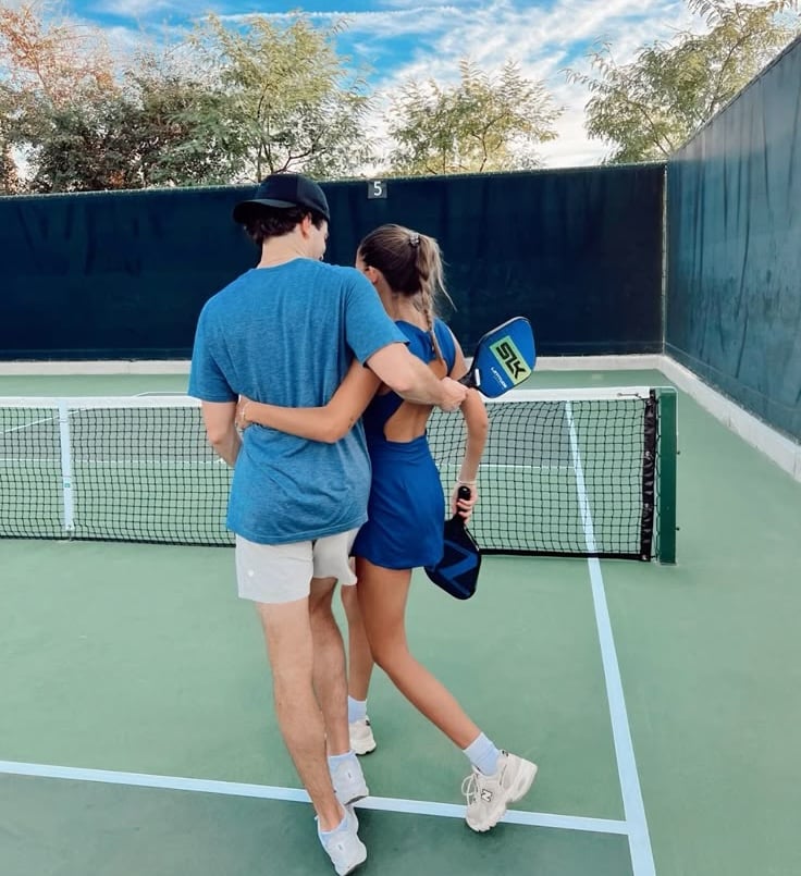 A young couple stands on a tennis court with their arms around each other, facing away from the camera. The woman holds a tennis paddle, and both are dressed in athletic clothing and sneakers.