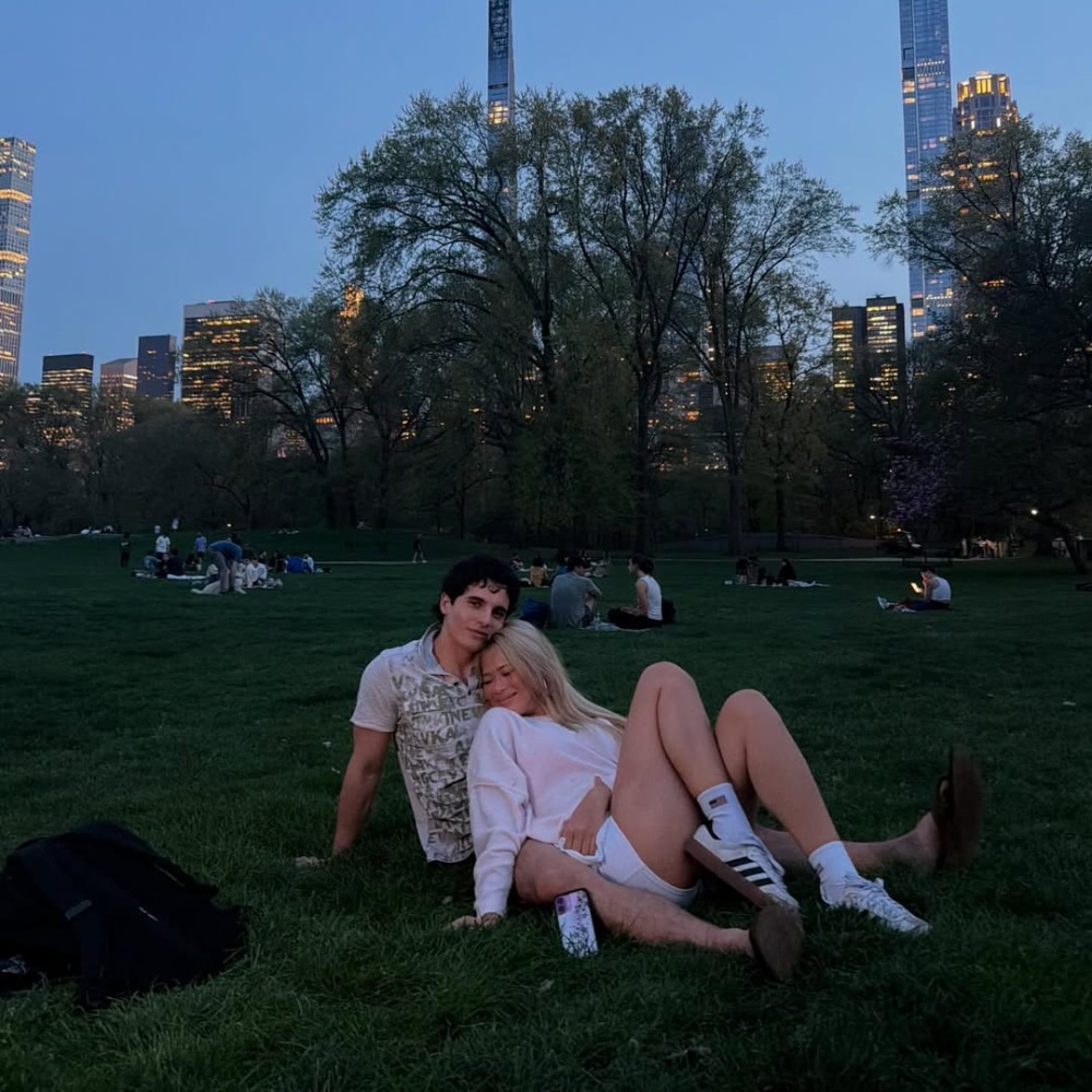 A young couple sits on grass in a park at dusk, sweetly flirting. The woman leans against her crush with her legs outstretched. City skyscrapers and trees rise in the background, while people are scattered around.