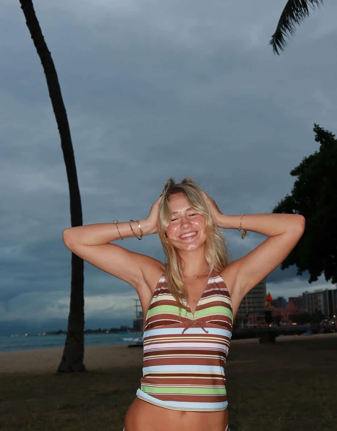 A young woman in a striped halter top smiles with her hands on her head at a beach near sunset, showcasing summer outfits amid palm trees, cloudy skies, and distant buildings in the background.