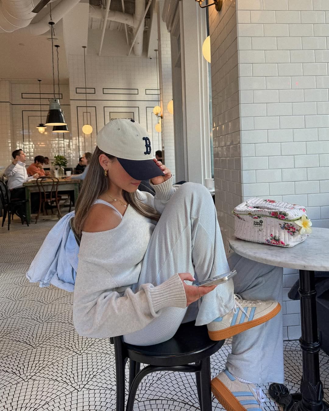 A woman in light loungewear and a baseball cap, perfect for summer outfits, sits sideways on a café chair, looking at her phone. She’s next to a small table with a floral bag. The café has white tiles, patterned floor, and a relaxed atmosphere.