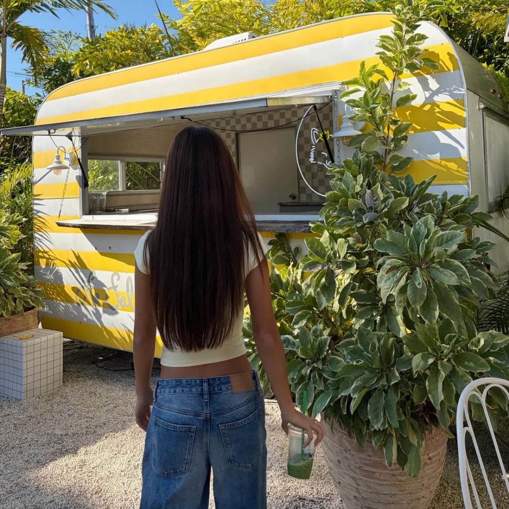A woman with long brown hair, wearing a yellow top and jeans, stands facing a yellow and white food truck surrounded by lush green plants—embracing cool girl summer vibes as she holds a green drink in her right hand.