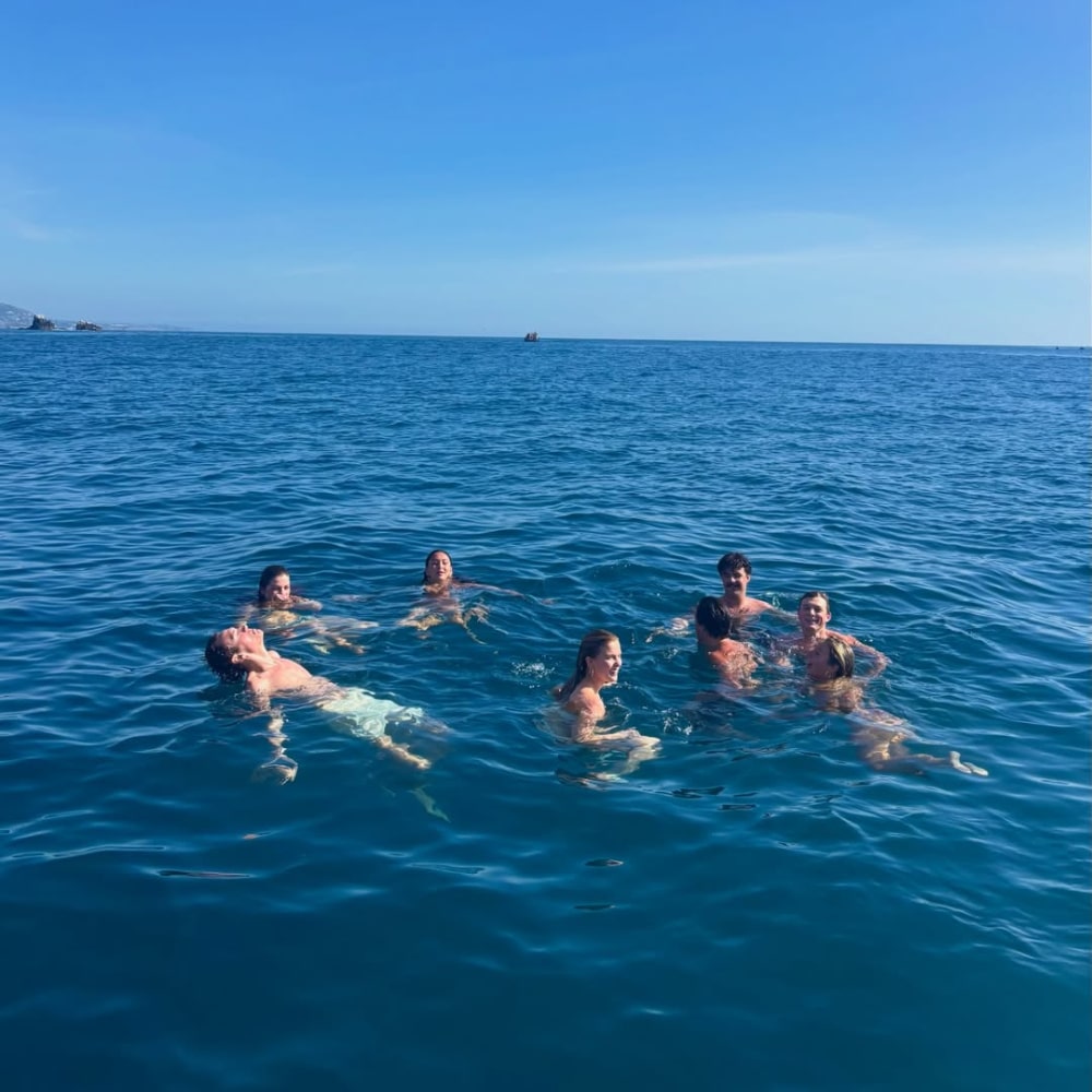 Seven people are swimming and floating together in a calm, blue sea under a clear sky, enjoying a cool girl summer on a budget. The water stretches to the horizon, creating a peaceful and relaxing scene.