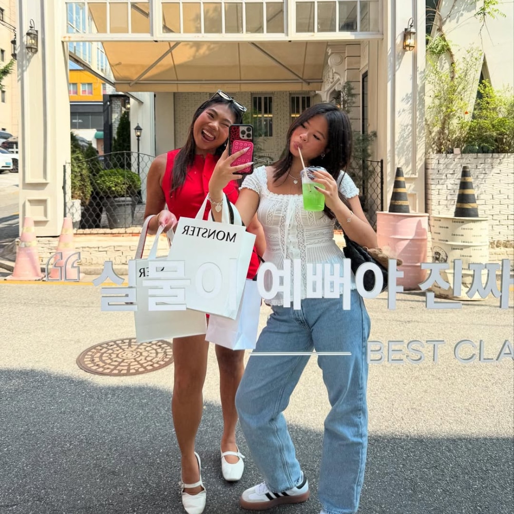 Two young women pose and smile outdoors in front of a glass door, capturing the essence of cool girl summer. One rocks a red dress and white shopping bag for a selfie, while the other sports a black top, jeans, and holds a green drink.