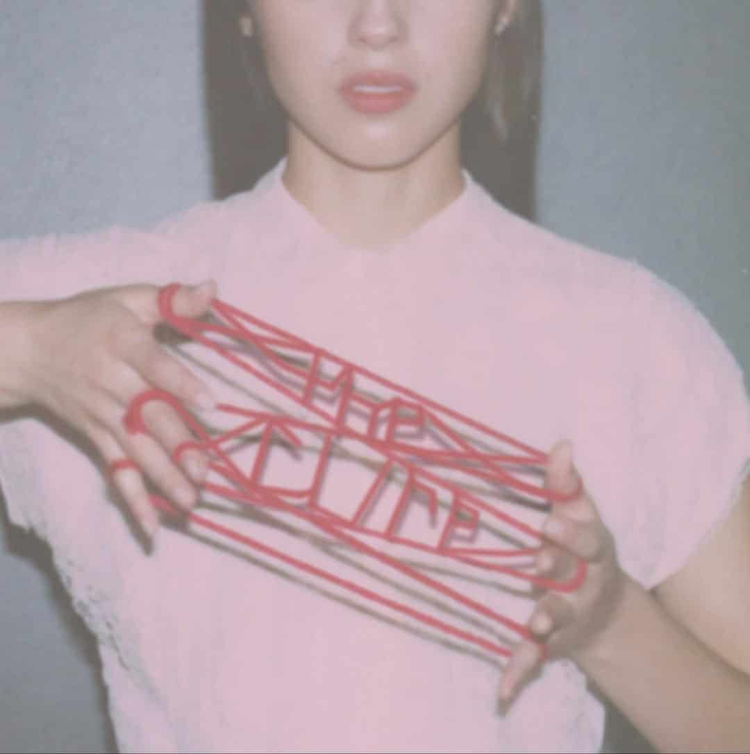 A person holds red string woven between their fingers in a cat’s cradle pattern, forming the word “HUMAN.” Channeling an Olivia Rodrigo vibe, they wear a white top and are shown from the shoulders up against a plain background.