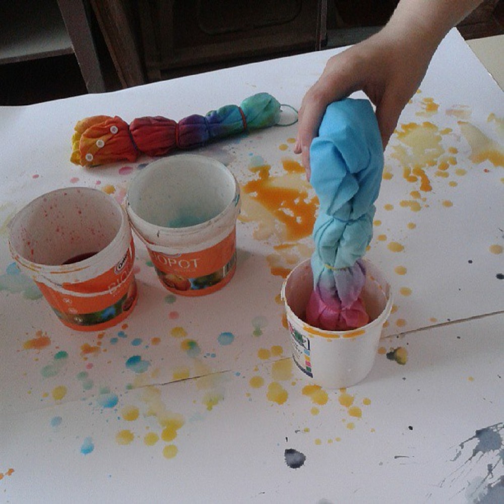 A hand dips a twisted, rubber-banded old clothes fabric into a cup of dye on a splattered table with two other dye containers, creating a vibrant tie-dye makeover.