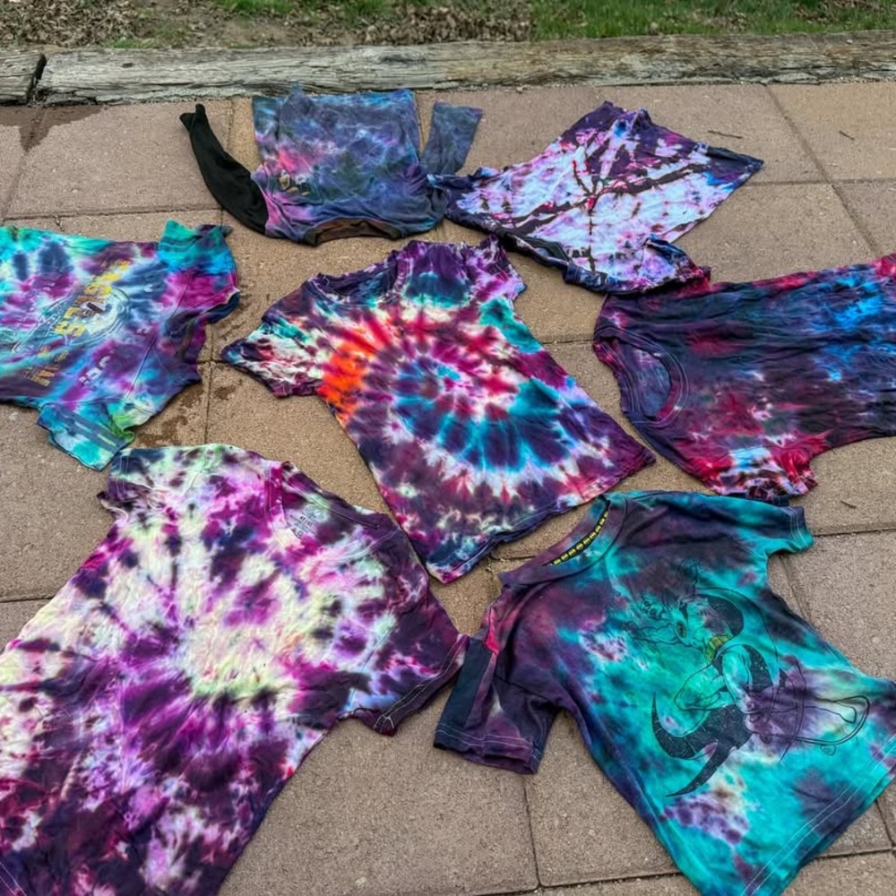 Seven colorful tie-dye t-shirts, made from old clothes in a fun makeover, are laid out on a paved surface with grass and wood in the background. The shirts display vibrant swirls and bursts of purple, blue, red, and more.