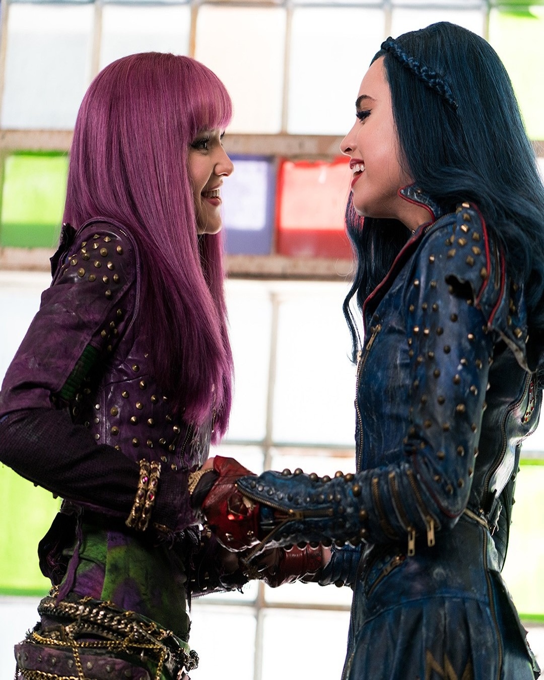Two young women in colorful, studded costumes stand facing each other, smiling and holding hands. One has long purple hair and the other has long blue hair. They are in front of a window with multicolored glass panes.