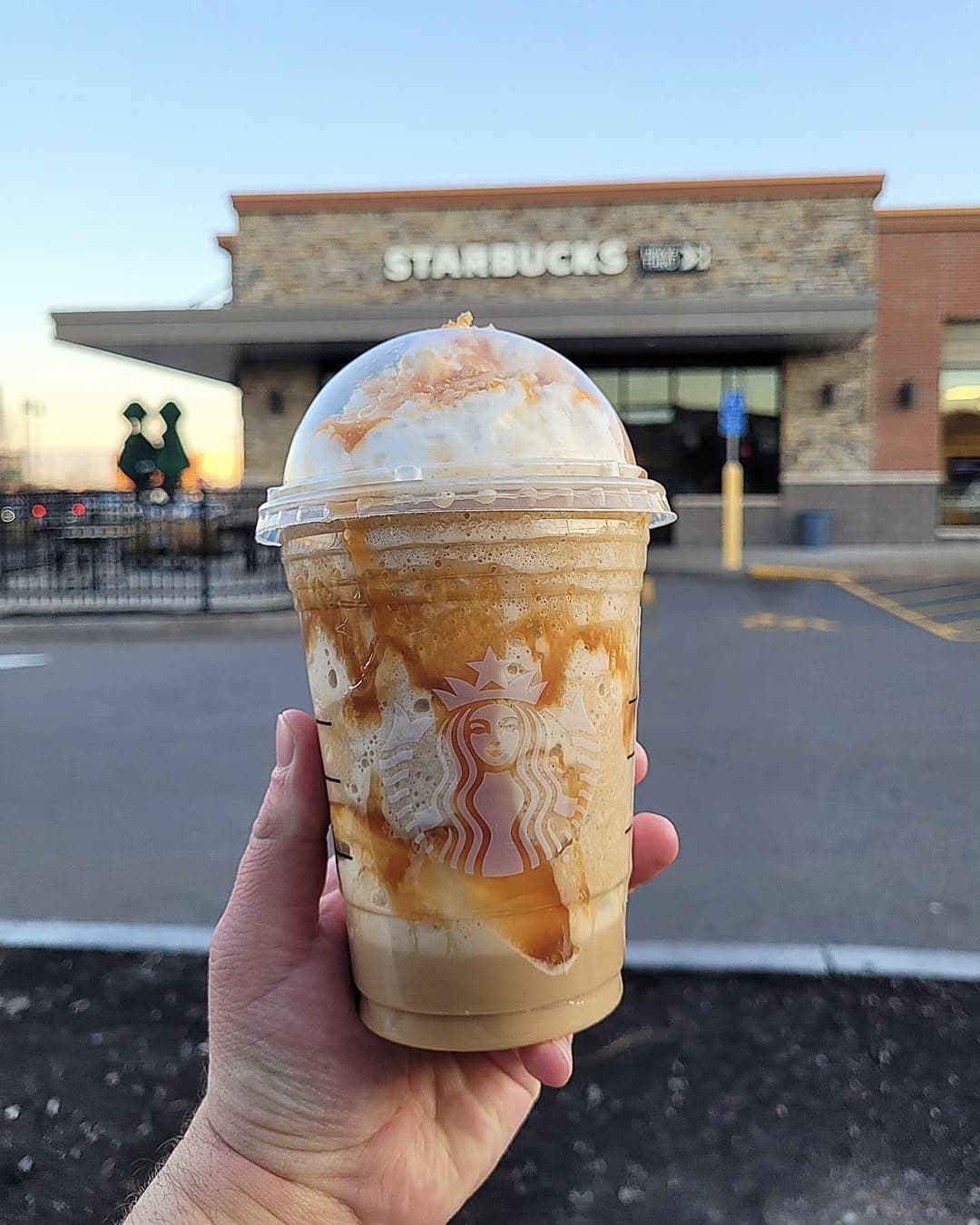 A hand holds a Starbucks caramel frappuccino topped with whipped cream and caramel drizzle in front of a Starbucks store on a clear day—perfect inspiration for recreating your favorite Starbies drinks at home.