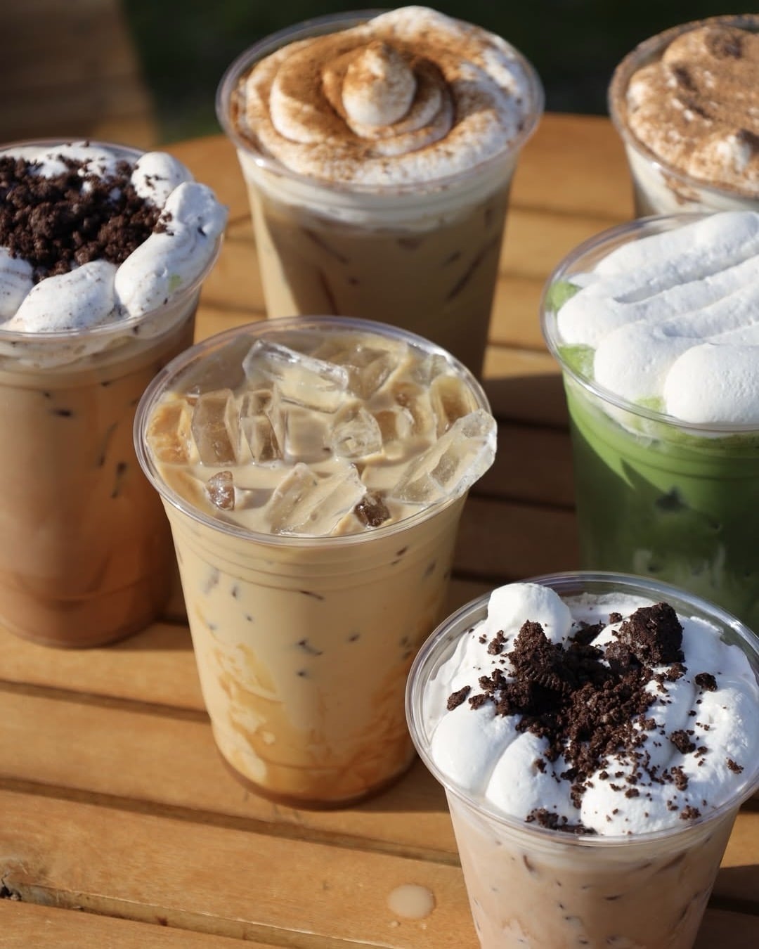 Six plastic cups of iced Starbucks drinks topped with whipped cream and various garnishes, including chocolate crumbs, cinnamon, and marshmallows, are arranged on a wooden table for a fun DIY treat at home.