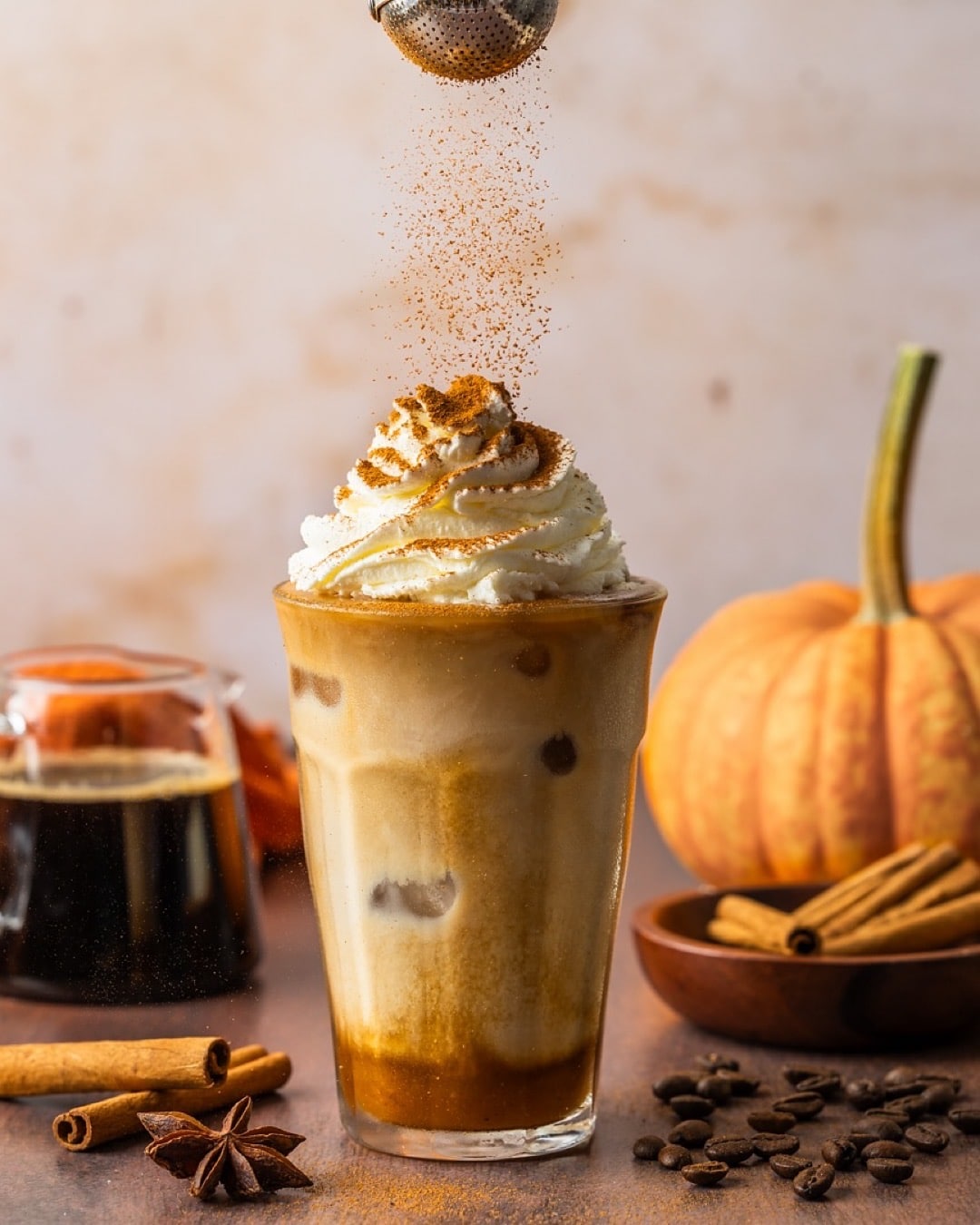 A glass of iced pumpkin spice latte topped with whipped cream and cinnamon, surrounded by a pumpkin, cinnamon sticks, star anise, coffee beans, and a small pitcher of coffee—a perfect DIY Starbucks drink to enjoy at home.
