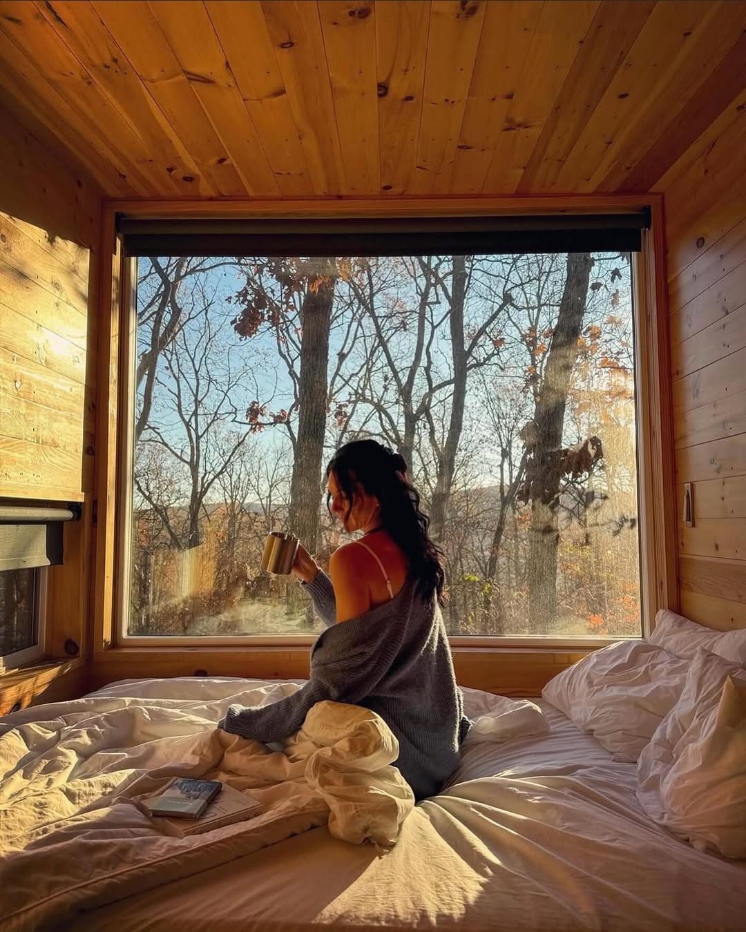 A woman with dark hair sits on a bed in a cozy wooden cabin, holding a mug and looking out a large window at bare trees and sunlight outside. A book rests on the unmade white bedding beside her.