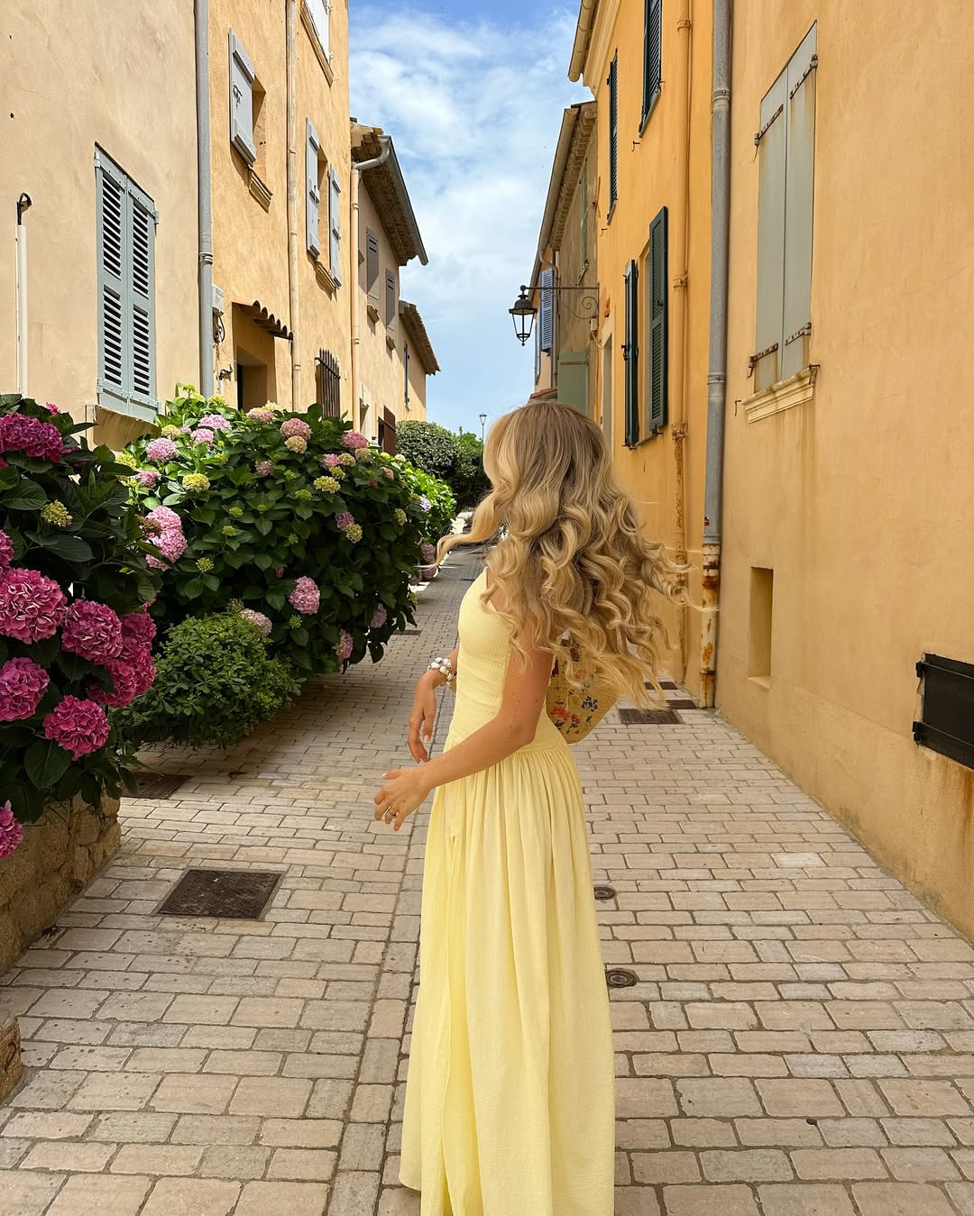 A woman with long, wavy blonde hair in a flowing yellow dress walks down a narrow, sunlit street lined with yellow buildings and blooming pink hydrangeas.