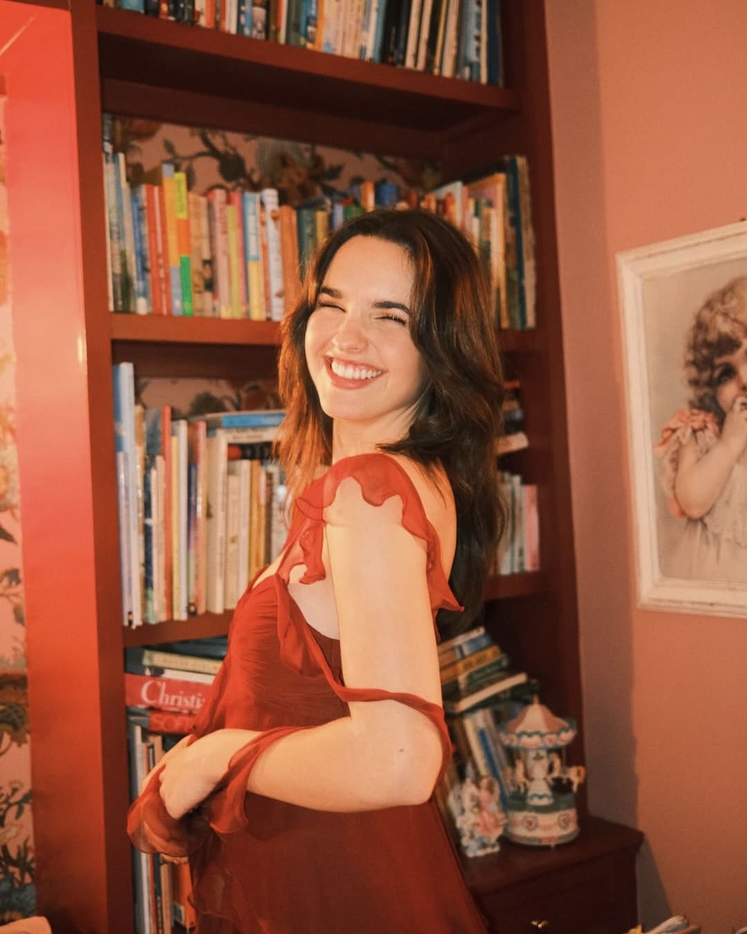 A young woman in a red dress smiles brightly while standing in front of a bookshelf filled with books. The room has pink walls and a framed picture of a cherub is hanging on the wall beside her.